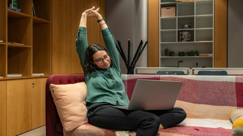 Mulher jovem de óculos e blusa verde sentada em um sofá moderno, sorrindo e se alongando com os braços para cima, enquanto usa um notebook em um ambiente de home office ergonômico.